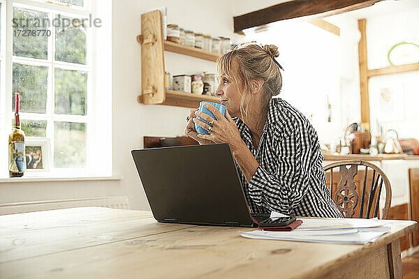 Lächelnde ältere Frau  die wegschaut  während sie Kaffee trinkt und mit einem Laptop in der Küche zu Hause sitzt