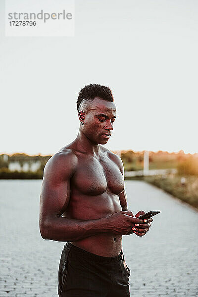 Junger männlicher Sportler  der bei Sonnenuntergang im Park steht und eine SMS auf seinem Smartphone schreibt