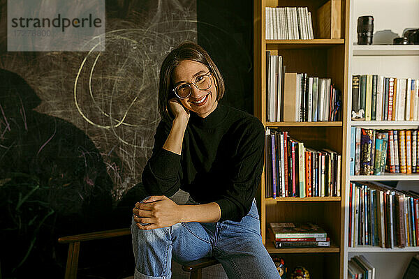 Lächelnde Frau mit Hand im Haar  die zu Hause am Bücherregal sitzt