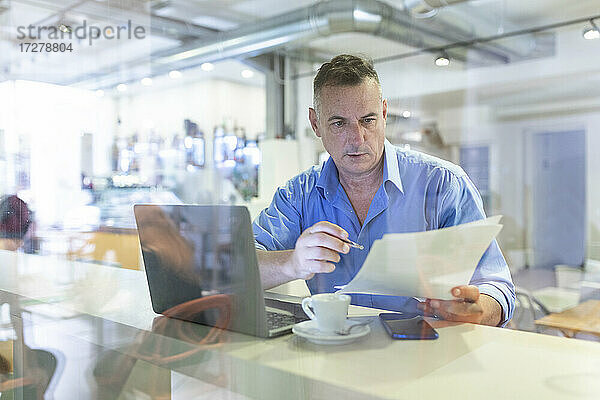 Männlicher Berufstätiger betrachtet ein Dokument  während er mit Laptop und Kaffee in einem Café sitzt