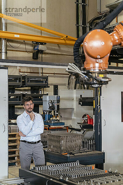 Geschäftsmann  der in einer Fabrik stehend eine Roboterarm-Maschine überwacht