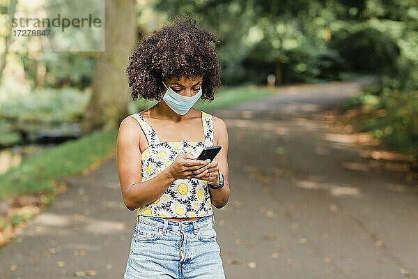 Mittlere erwachsene Frau mit Gesichtsmaske  die ein Mobiltelefon benutzt  während sie auf der Straße im Park steht