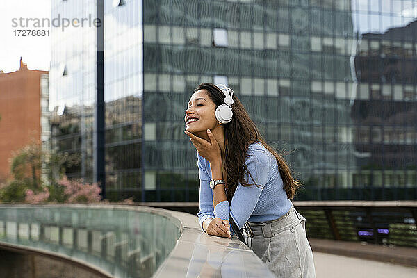 Lächelnde Geschäftsfrau  die Musik über Kopfhörer hört  während sie sich an das Geländer einer Fußgängerbrücke lehnt