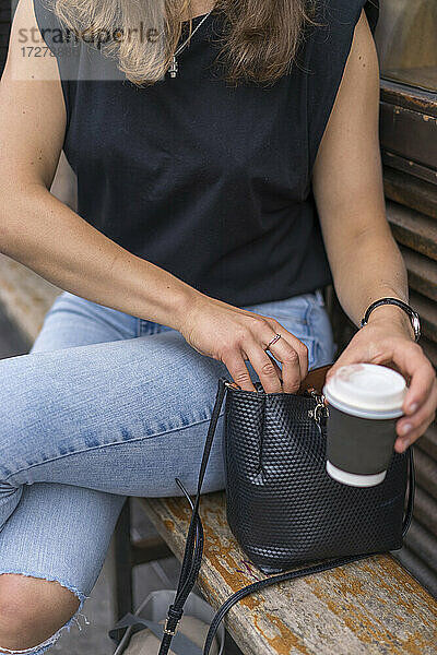 Frau  die ihre Hand in die Handtasche steckt und einen Einweg-Kaffeebecher hält  sitzt auf einer Bank