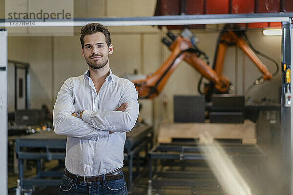 Junger Geschäftsmann steht mit verschränkten Armen vor einem Roboterarm in einer Fabrik