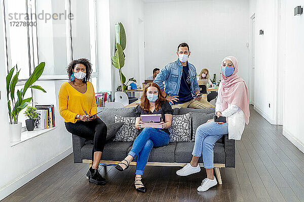 Mitarbeiter mit Gesichtsmaske  die soziale Distanz wahren  während sie mit einem Kollegen im Hintergrund im Büro sitzen  während COVID-19