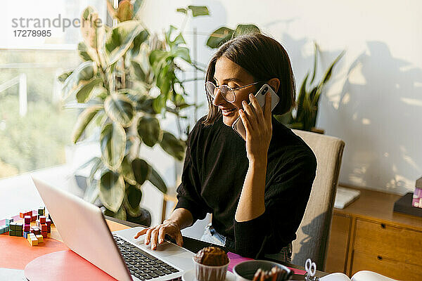 Mittlere erwachsene Geschäftsfrau  die mit einem Smartphone telefoniert  während sie einen Laptop benutzt und zu Hause sitzt