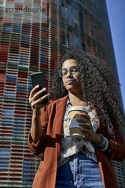 Frau  die ein Smartphone benutzt und einen Bambusbecher hält  während sie an einem Gebäude in der Stadt steht