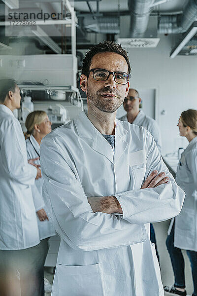 Männlicher Wissenschaftler steht mit verschränkten Armen und einem Mitarbeiter im Hintergrund in einem Labor