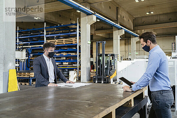Geschäftsmann mit Gesichtsmaske  der ein digitales Tablet benutzt  während er in sozialer Distanz zu einem Kollegen in einer Fabrik steht