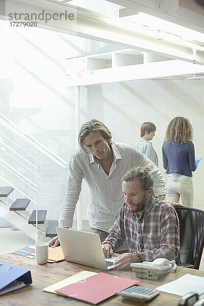 Mann arbeitet an einem Laptop mit einer Mitarbeiterin im Hintergrund im Büro