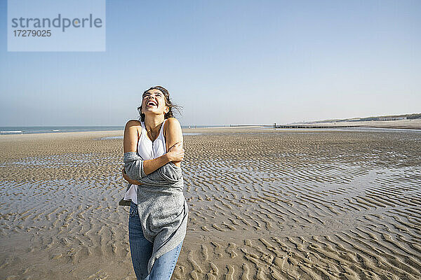 Glückliche junge Frau  die sich selbst umarmt  während sie am Strand gegen den klaren Himmel steht