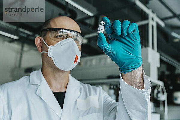 Wissenschaftler mit Schutzmaske und Brille  der eine Impfstoffflasche hält  während er im Labor steht