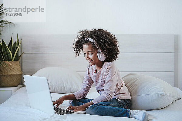 Nettes Mädchen  das einen Laptop benutzt und Kopfhörer trägt  während es im Schlafzimmer zu Hause sitzt