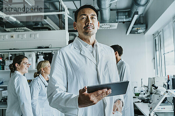 Wissenschaftlerin mit Hand in der Tasche und digitalem Tablet  stehend mit Mitarbeitern im Hintergrund im Labor