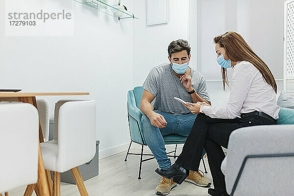 Arzt und Patient tragen eine Gesichtsmaske und benutzen ein digitales Tablet  während sie im Büro sitzen