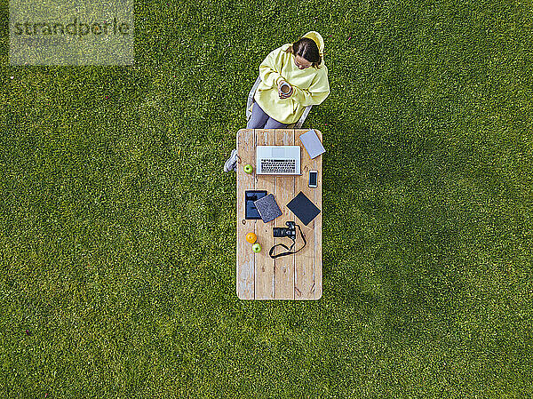 Luftaufnahme einer Frau  die an einem Kaffeetisch auf grünem Rasen sitzt und eine Tasse Kaffee in den Händen hält