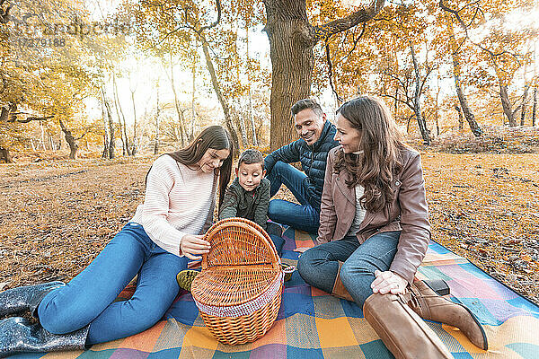 Kinder beim Öffnen eines Essenskorbs  während die Eltern im Herbst auf einer Picknickdecke im Park sitzen