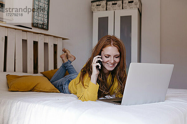 Lächelnde Frau  die auf der Vorderseite liegt und mit ihrem Smartphone im Schlafzimmer telefoniert