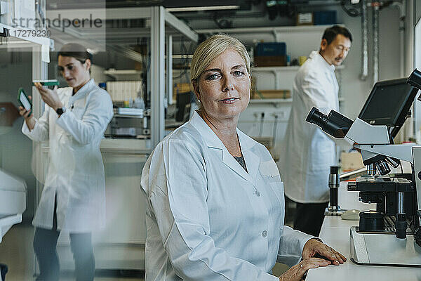 Wissenschaftlerin am Mikroskop sitzend mit einem Mitarbeiter im Hintergrund  der in einem Labor arbeitet