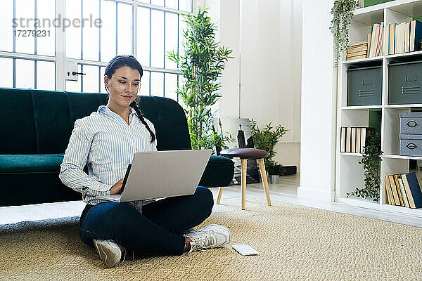 Junge Frau  die einen Laptop benutzt und mit gekreuzten Beinen auf dem Boden vor dem Sofa sitzt