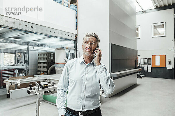 Männlicher Ingenieur schaut weg  während er in einer Fabrik mit seinem Smartphone telefoniert