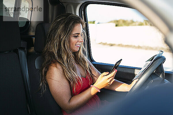 Lächelnde Frau mit Landkarte  die ein Mobiltelefon benutzt  während sie im Wohnmobil am Strand sitzt