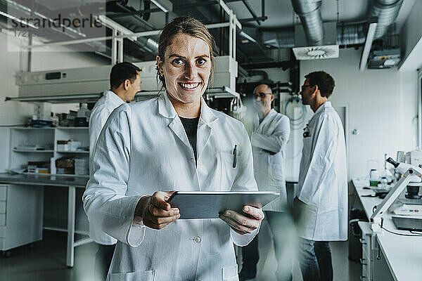 Lächelnde Frau  die ein digitales Tablet benutzt  während sie mit einem Mitarbeiter im Hintergrund in einem Labor steht