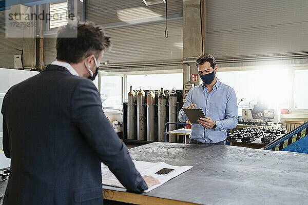 Junger Geschäftsmann mit digitalem Tablet und Gesichtsmaske bei der Arbeit  während er mit einem Kollegen in sozialer Distanz in einer Fabrik steht