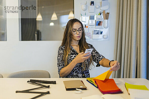 Kreative Geschäftsfrau  die ein Stoffmuster mit ihrem Smartphone fotografiert  während sie am Schreibtisch im Büro sitzt