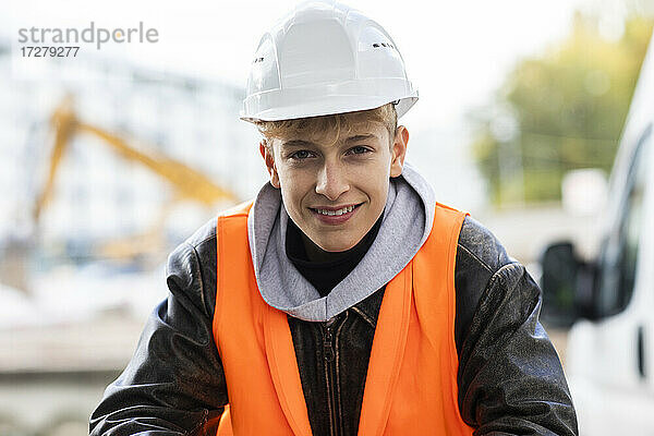 Lächelnder jugendlicher Baupraktikant mit Schutzhelm und reflektierender Kleidung auf der Baustelle