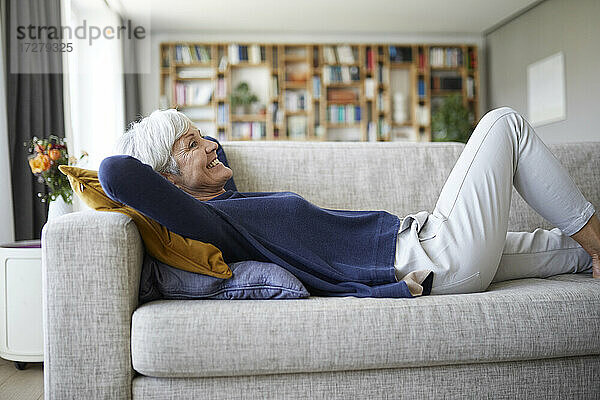 Lächelnde ältere Frau mit Händen hinter dem Kopf  die zu Hause auf dem Sofa liegt