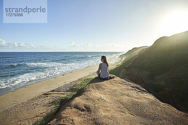 Weibliche Touristin sitzt auf einer Felsformation und schaut aufs Meer