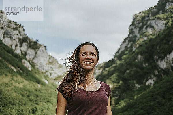 Lächelnde Wanderin mit langen braunen Haaren  die wegschaut  während sie vor einer Bergkette steht