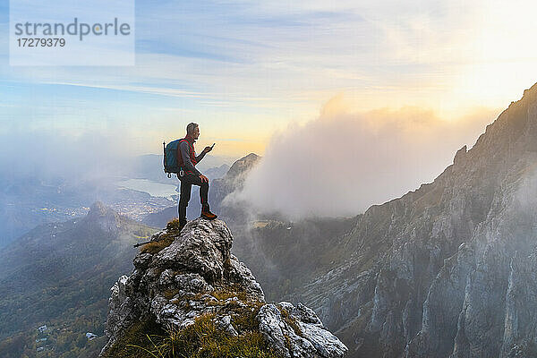 Nachdenklicher Wanderer mit Smartphone auf einem Berggipfel bei Sonnenaufgang in den Bergamasker Alpen  Italien