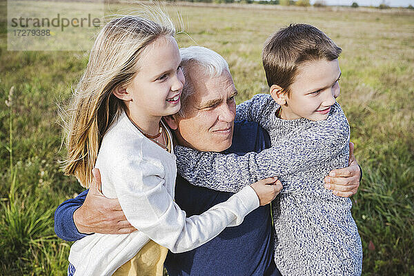 Großvater schaut weg  während er seine süßen Enkelkinder auf dem Feld umarmt