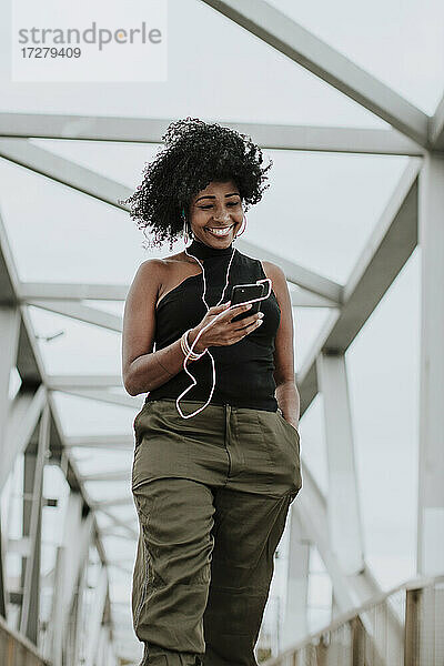 Lächelnde Frau  die ein Smartphone benutzt und über Kopfhörer Musik hört  steht auf einer Brücke in der Stadt