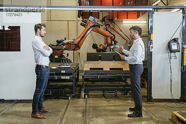 Geschäftsmann im Gespräch mit einem Kollegen  während er in einer Fabrik vor einem Roboterarm steht