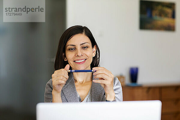 Lächelnde Frau  die auf einen Stift schaut  während sie einen Laptop zu Hause benutzt