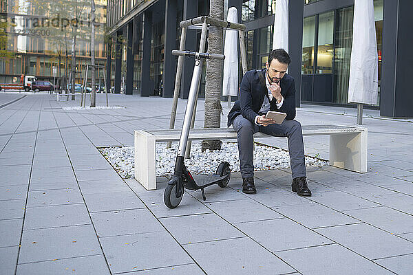 Geschäftsmann  der ein digitales Tablet benutzt  während er mit einem E-Scooter auf einer Bank in der Stadt sitzt