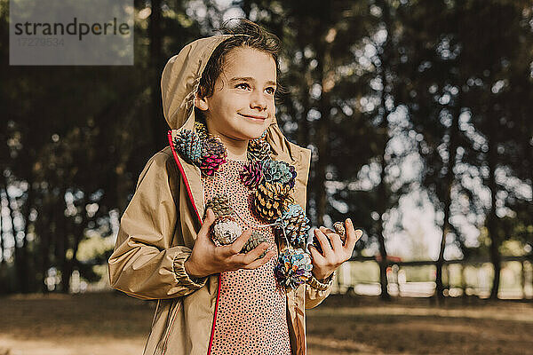 Lächelndes niedliches Mädchen mit bunter Tannenzapfengirlande schaut weg  während sie im Park steht