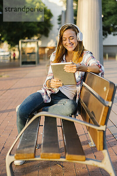 Lächelnde Frau  die ein digitales Tablet benutzt  während sie an einem sonnigen Tag auf einer Bank sitzt