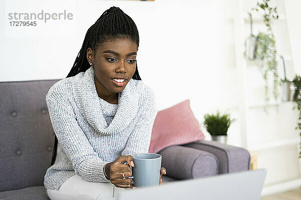 Junge Unternehmerin hält eine Kaffeetasse  während sie zu Hause am Laptop arbeitet