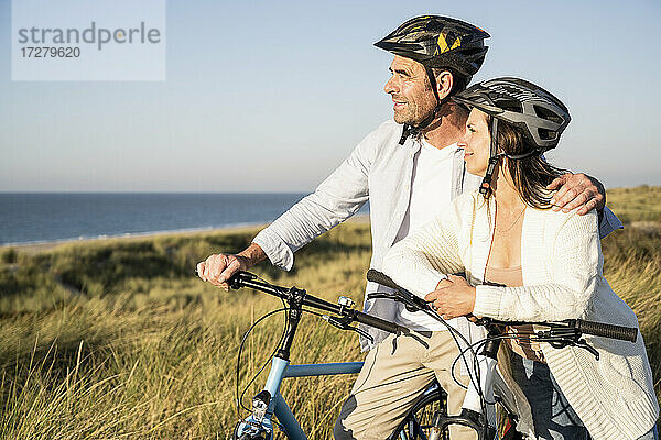 Lächelndes reifes Paar  das die Aussicht betrachtet  während es mit Fahrrädern am Strand steht