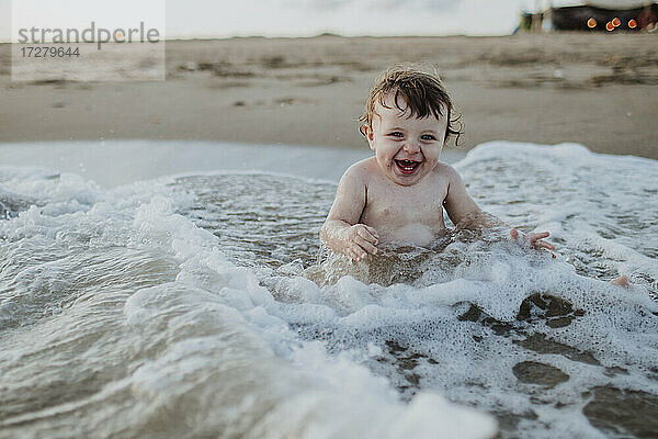 Männliches Kleinkind lachend im Wasser sitzend am Strand bei Sonnenuntergang