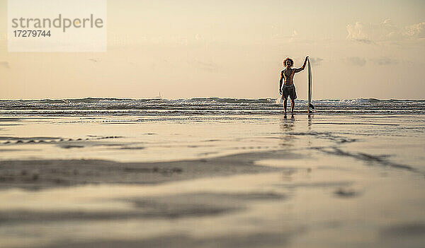 Mann ohne Hemd steht mit Surfbrett am Meer gegen den Himmel bei Sonnenuntergang