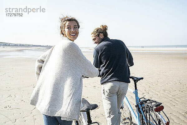 Lächelnde junge Frau  die mit Fahrrad und Freund spazieren geht  während sie über die Schulter auf den Strand gegen den klaren Himmel an einem sonnigen Tag schaut