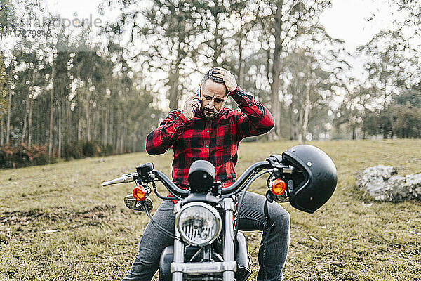 Angespannter männlicher Motorradfahrer  der auf einem Motorrad im Wald sitzt und mit seinem Smartphone telefoniert