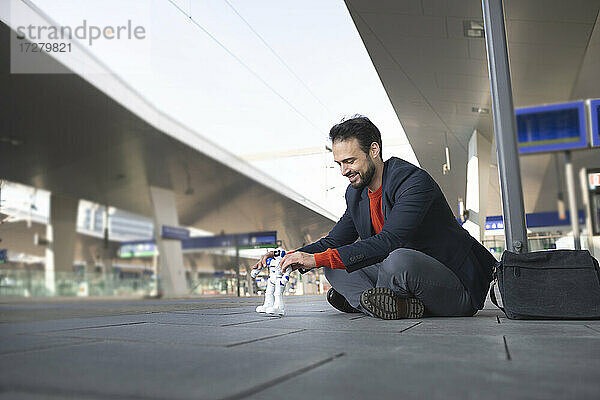 Lächelnder Unternehmer  der einen Roboter betrachtet  während er auf einem Bahnsteig sitzt