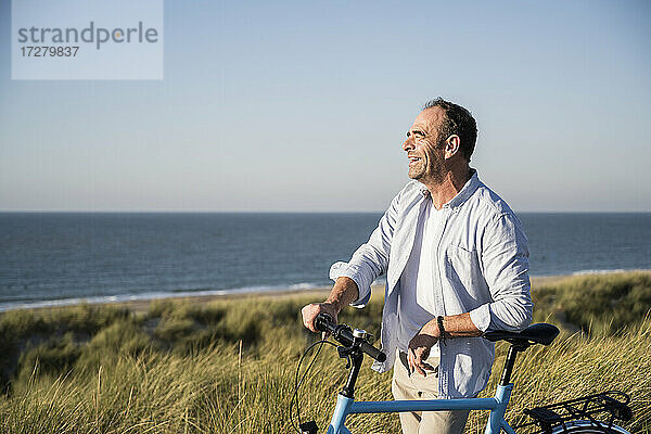 Fröhlicher reifer Mann  der wegschaut  während er mit dem Fahrrad am Strand vor dem klaren Himmel steht